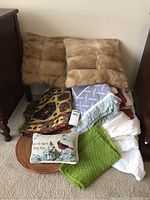 Grouped items including two beige faux fur pillows, patterned table runners, green knitted cloth, embroidered small pillow and folded white linen.