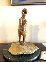 Front view of the signed sculpture showing pregnant figure standing on a rough light-colored stone base placed on a circular dark granite base.