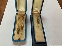 Opened blue velvet-lined boxes showing three decorative stickpin heads and two screw-on backings