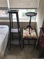 Photo showing tall black wooden plant stand, floral upholstered footstool, and small tray table near window with light carpet floor.