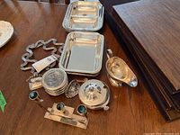 Photo of two rectangular silver-plated serving trays, silver-plated gravy boat, copper plate butter dish with lid, silver napkin ring, assorted small silver pieces, and a decorative item possibly a coaster holder on a wooden table.