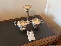 Photo showing sterling silver candlestick alongside creamer and sugar bowls on a dark surface.
