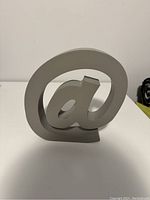 Front view of gray '@' symbol decorative desk ornament on white surface.