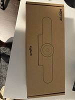 Top view of the unopened rectangular brown cardboard box with black line sketch of the Logitech Meetup camera and brand text.