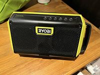 Front view of Ryobi portable speaker showing black grille and yellow side panels with Ryobi logo.