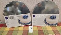 Two Wilton 14 inch cake platters in box on table with checkered cloth, showing front of box with product image and name
