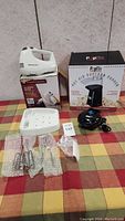 Three kitchen appliances on a checkered cloth, including a white Toastmaster hand mixer on its box, Popflix hot air popcorn popper in box, and a black electric can opener.