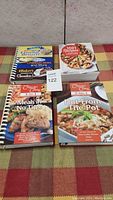 Photo showing four cook books laid flat on a checkered fabric surface. Titles visible: Favorite Desserts and More, 1001 Recipes, Meals in No Time, Hot From The Pot.