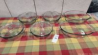 Full lot photo showing all six transparent glass Anchor bakeware dishes with lids arranged on a tablecloth with a checkered pattern.