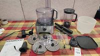 Full set of Kenwood food processor and blender attachments laid out on table including base unit, bowl, discs, pusher, blender jug, and manual.