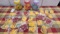 Wide shot showing an assortment of yellow, red, and other colored cookie cutters in plastic bags spread on a table, with three jars containing more cookie cutters in the background