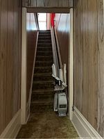 Photo of a straight stair chair lift installed along a wooden-paneled staircase. The chair lift is mounted on a rail on the right side of the stairs, designed to travel the length of 7 carpeted steps.