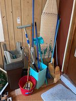Multiple brooms, mop, squeegee, blue and red buckets, cleaning caddy, and ironing board visible in corner near wooden wall paneling