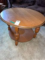 Round oak coffee table showing top and carved legs with a lower shelf, sitting on carpet next to dark fabric sofa.