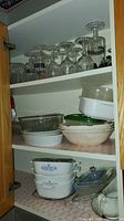Kitchen cabinet shelves with various glass items: stemware glasses on top shelf, ceramic and glass bowls in middle and bottom shelves, including CorningWare dishes and glass containers.
