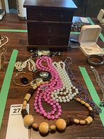 Photo showing the wooden jewelry box in the background with multiple necklaces and bracelets spread out in front, including pearl-like necklace, pink and purple bead necklaces, wooden bead necklace and a floral brooch.