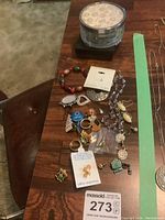 View of assorted costume jewelry items including pins, rings, earrings, bracelet, and a round glass vanity box on wooden table.