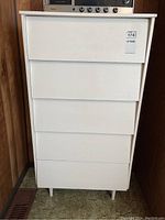 Front view of white five drawer dresser showing the drawer fronts and legs against wood panel wall and green carpet