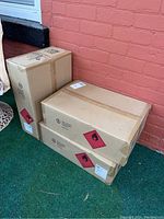 Three sealed cardboard boxes stacked near a red brick wall, each box labeled for hand sanitizer.