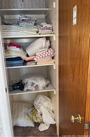 Closet view showing multiple folded towels, sheets, and pillowcases along with hair tools in plastic bag on shelf.