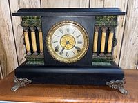 Front view of a vintage mantle clock with Roman numeral clock face, ornate gold bezel, black casing with green and yellow faux marble accents, standing on decorative metal feet.