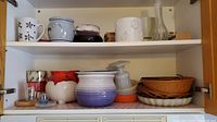 Shelf showing multiple ceramic mugs, pots, bud vase, wooden nutcracker and dishes.