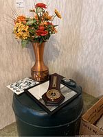 Copper-tone vase holding faux flowers, wooden barometer thermometer, tile art, photograph, and round green ottoman arranged together