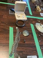Photo of multiple silver jewelry items including a filigree bracelet in white box, oxidized bangle, circular brooch/pendant, ring with black stone, and chain bracelets on wood surface.