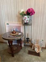 Wooden round table with glass top, metal plant stand with floral bucket and faux flowers, framed print leaning against wall, crystal candlesticks, and decorative items on floor