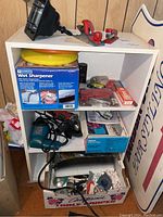 Front and side view of white shelving unit filled with boxed tools and miscellaneous hardware and household items including sharpener, angle grinder, cordless drill, and seashells box.