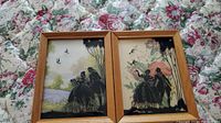 Two framed silhouette 3D wall art pieces placed side-by-side on a floral cloth background, showing detailed figures and scenery.