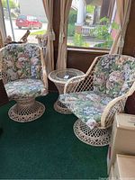 Photo shows two lacquered wicker swivel arm chairs with floral cushions alongside a round lacquered wicker side table with a glass top. Set placed indoors near windows with green carpet beneath.
