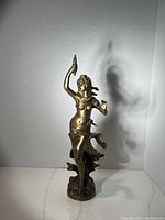 Front view of bronze statue showing female figure raising a hand with an object, draped in flowing fabric, on a base with floral details.