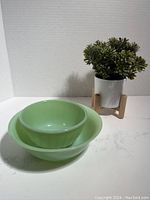 Two green Fire King Jadeite swirl mixing bowls displayed, one nested inside the other, on a white surface with a small plant background.