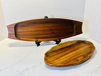Two teak wood charcuterie trays displayed on white surface, one large oval tray with handles on stand and smaller scalloped tray in front.