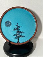 Full view of the circular ceramic plate on a black stand showing the vivid turquoise blue background with a black stylized fir tree and moon.