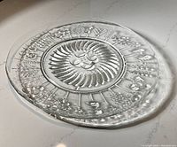 Top-down angled view of the round clear glass serving plate with detailed floral pattern and central swirl design, highlighting the embossed raised elements and nearly flat surface.