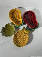 Four Olfaire Portugal majolica serving dishes: yellow pineapple, red eggplant, yellow eggplant, and green grape leaf, arranged on a white surface.