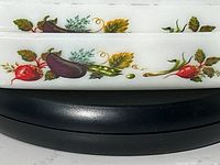 Close-up showing vegetable and herb pattern decoration on white glass casserole dishes with detailed colorful radishes, eggplants, and leaves.