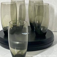 Five smoked gray hand blown hi ball glasses arranged on a circular black platform, showing their translucent tint and oval shape.