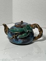 Side view of the blue pottery teapot with a vine shaped handle and pea pod decorative elements showing the colors and shape clearly.