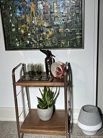 Full front view of the vintage chrome and laminated wood serving trolley beside a wall with colorful abstract painting. Shows two-tier shelving with plant on lower shelf and glassware and decorative items on top shelf.