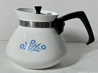 White teapot with Blue Cornflower floral design, black handle and knob, stainless steel collar around lid area.