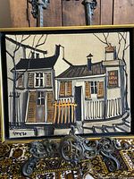 Framed oil on canvas painting showing two stylized houses with a sign 'AU PAIN AGILE', signature and number at bottom left, propped on an ornate metal stand on patterned fabric.