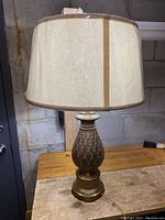 Full view of the extra large pineapple-shaped brass table lamp with beige fabric shade on a wooden surface in front of a concrete block wall.