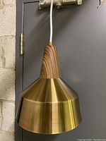 Front side view of MCM pendant lamp showing wooden top and brass shade hanging against a gray door and brick wall background.