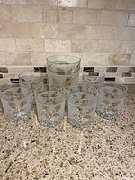 Seven pieces of cocktail glassware including six frosted tumblers and one matching frosted ice bucket, all decorated with gold leaf and vine designs, placed on countertop.