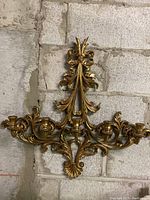 Front view of the gold tone metal wall hanging candle holder with five candle holders and detailed decorative scroll and floral motif.