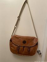 Front view of the Apansy vintage leather messenger bag hanging on a hook against a plain wall, showcasing the front flap with the logo patch, buckle straps and adjustable beige shoulder strap.