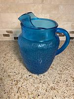 Front side view of vintage blue glass pitcher showing pagoda style rim and textured geometric pattern on body.